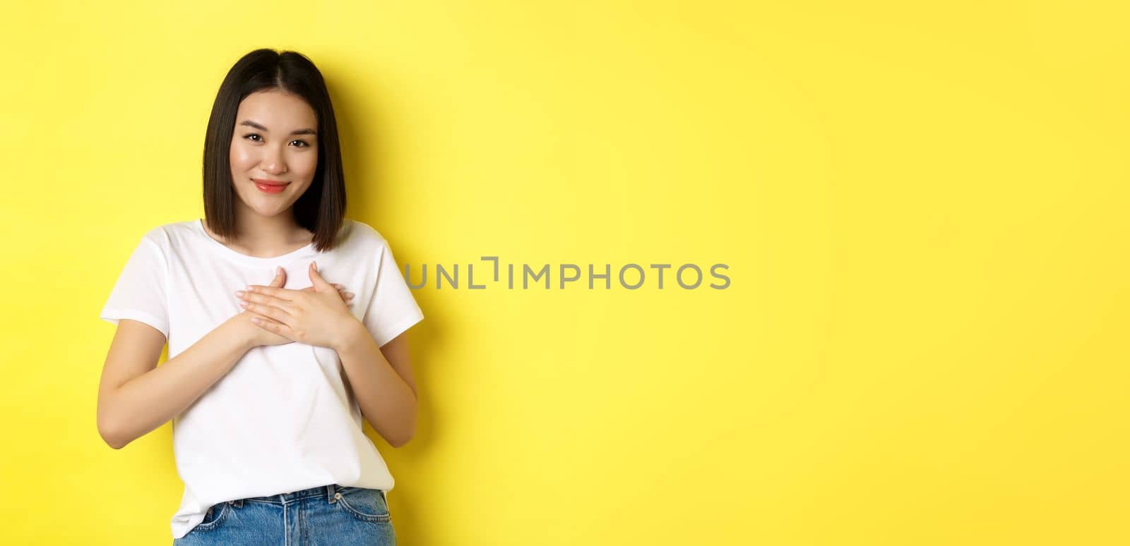 Beauty and fashion concept. Heartfelt asian girl holding hands on heart and smiling touched, thanking you, standing over yellow background.