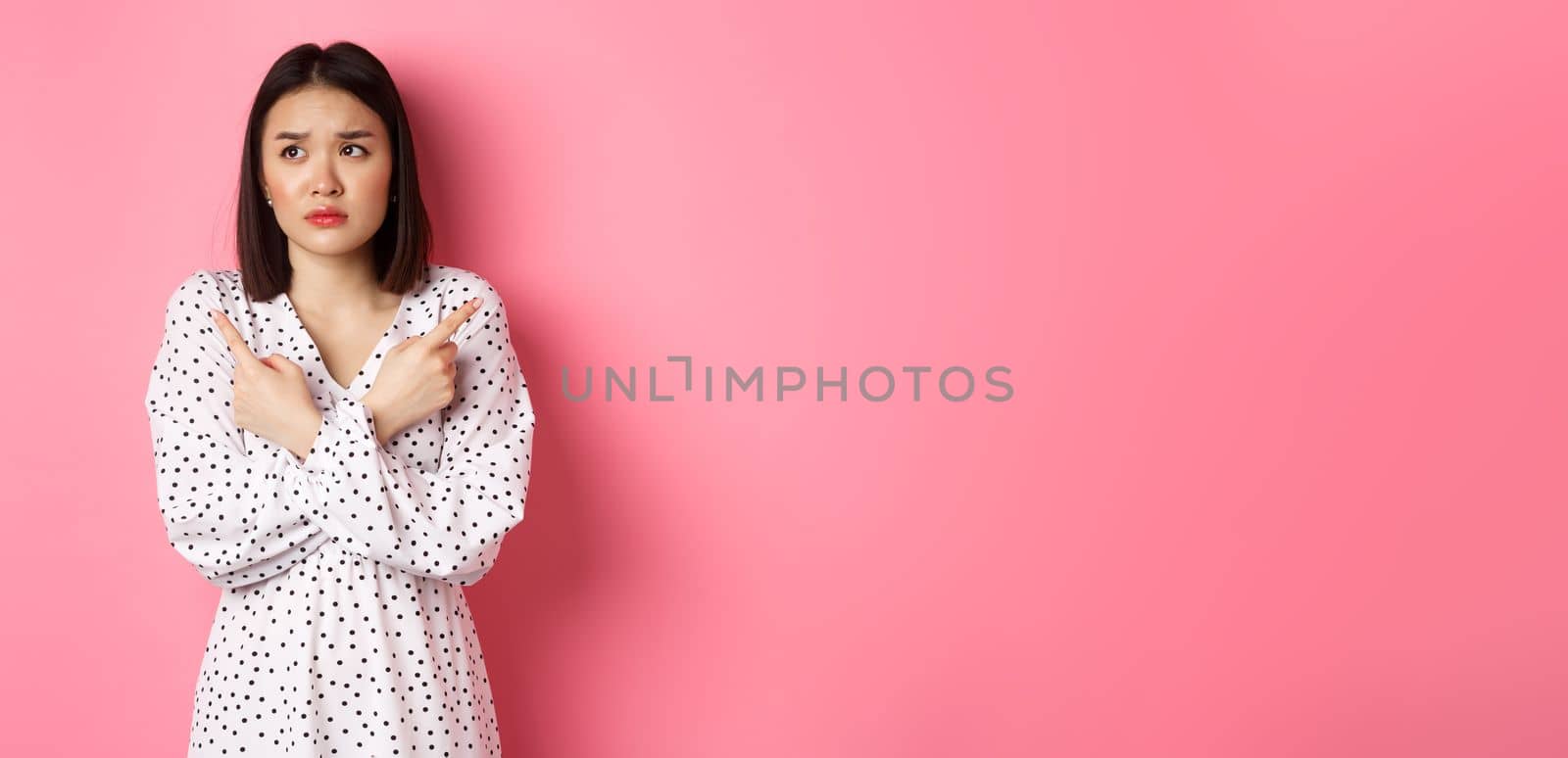 Concerned asian woman having doubts, pointing sideways and looking left with hesitant and sad face, need help with choice, standing over pink background.