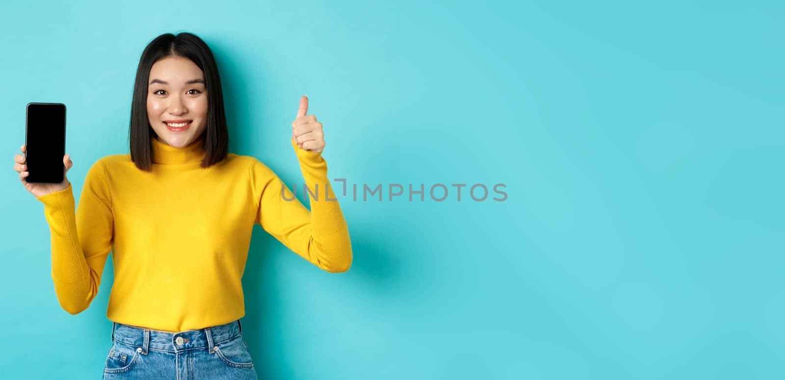 Technology and people concept. Cheerful asian girl in yellow sweater showing blank smartphone screen and thumbs up, demonstrate online offer, standing over blue background by Benzoix