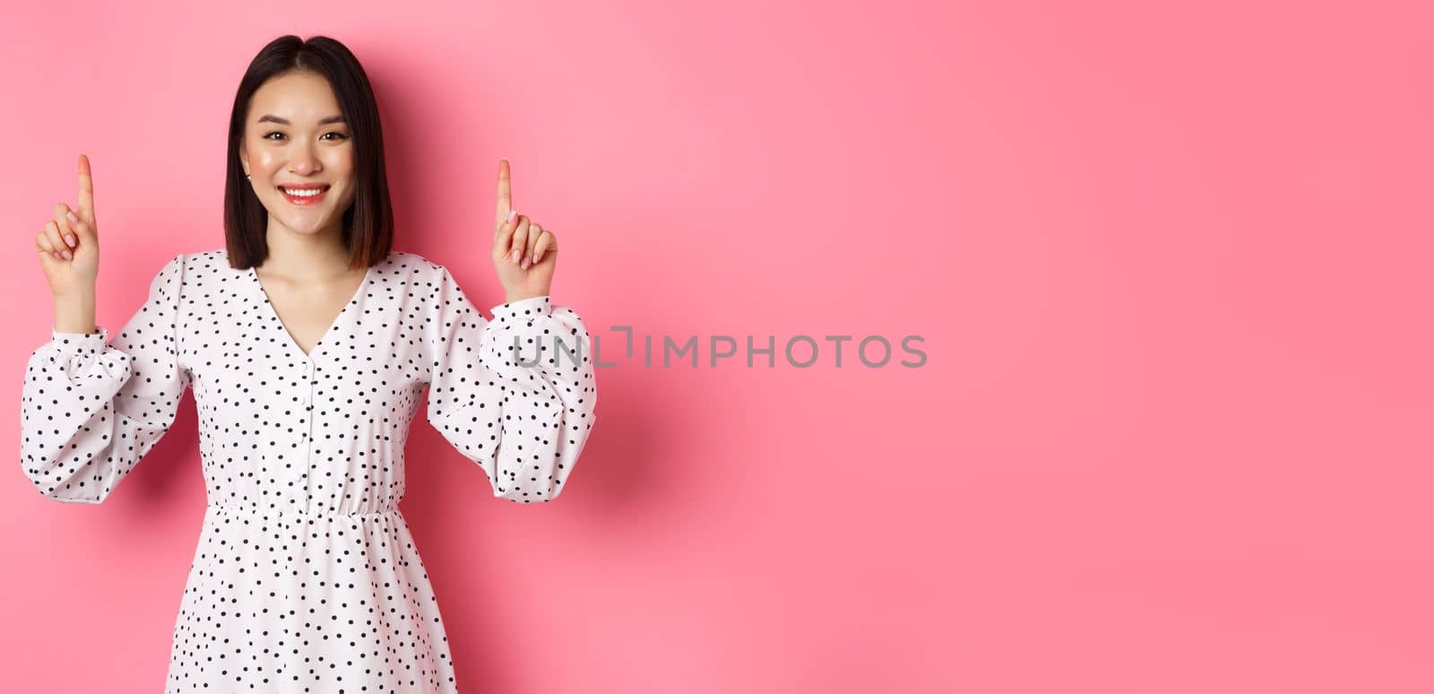 Beautiful Korean young woman in dress showing announcement, pointing fingers up at promo copy space, smiling happy, standing over pink background.
