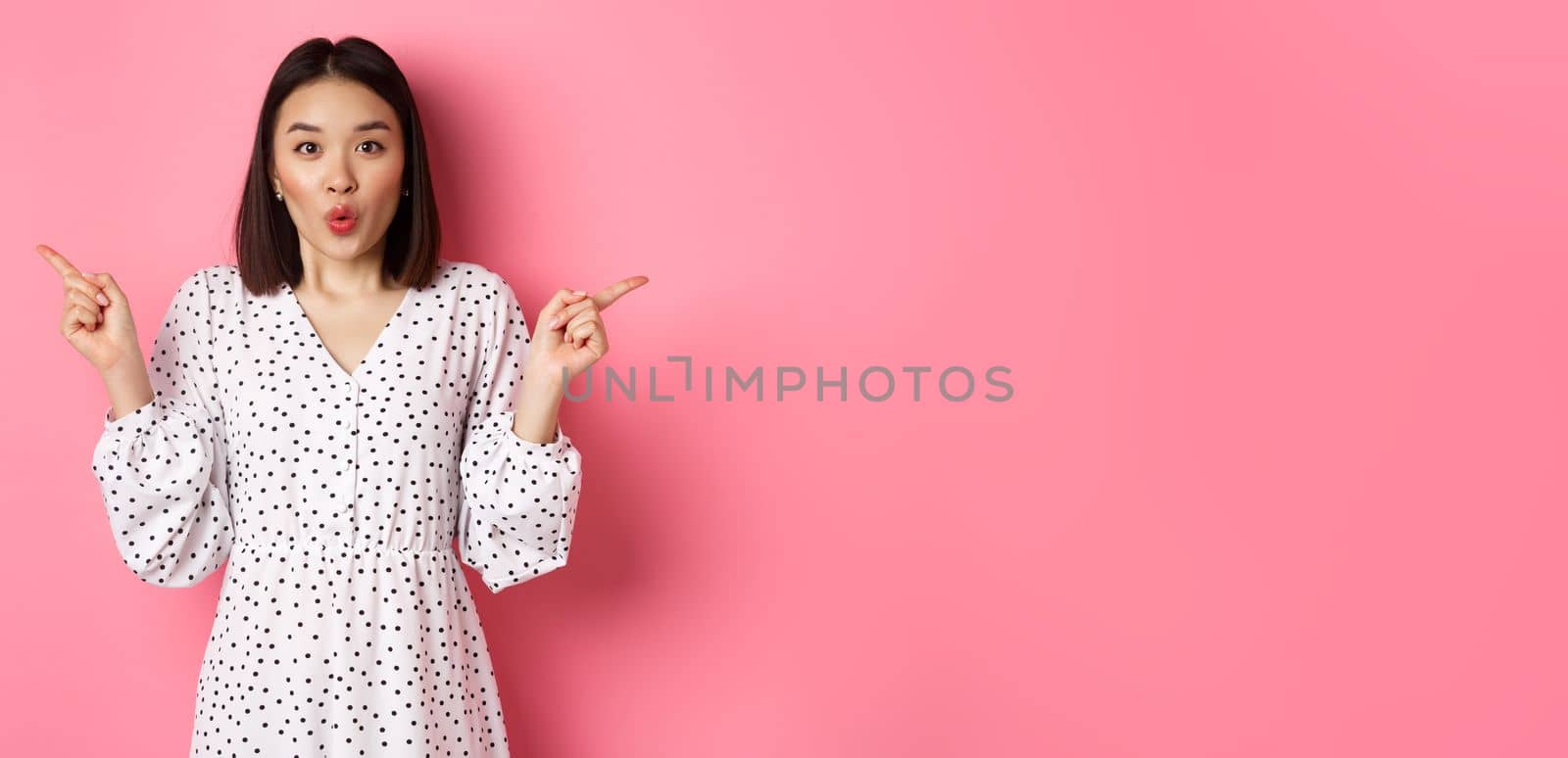 Beautiful asian woman making choice on shopping, pointing fingers sideways and showing variants, smiling at camera, standing over pink background.