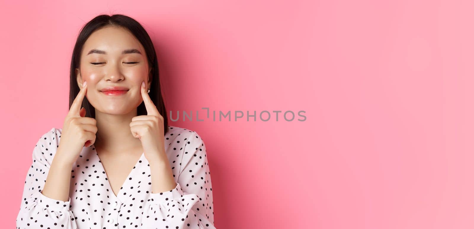 Beauty and lifestyle concept. Close-up of beautiful asian woman poking cheeks with closed eyes, smiling satisfied, standing over pink background.