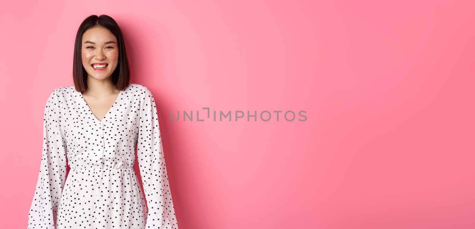 Happy korean woman in dress looking at camera, smiling and laughing with sincere expression, standing over pink background by Benzoix
