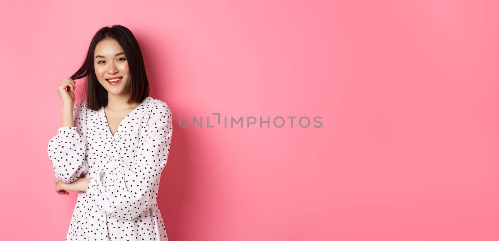 Cute asian female brunette in trendy dress, playing with hair and smiling flirty, standing in dress over pink background.