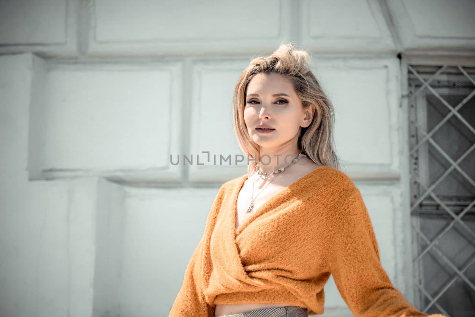 A middle-aged woman looks like a good blonde with curly beautiful hair and makeup on the background of the building. She is wearing a yellow sweater