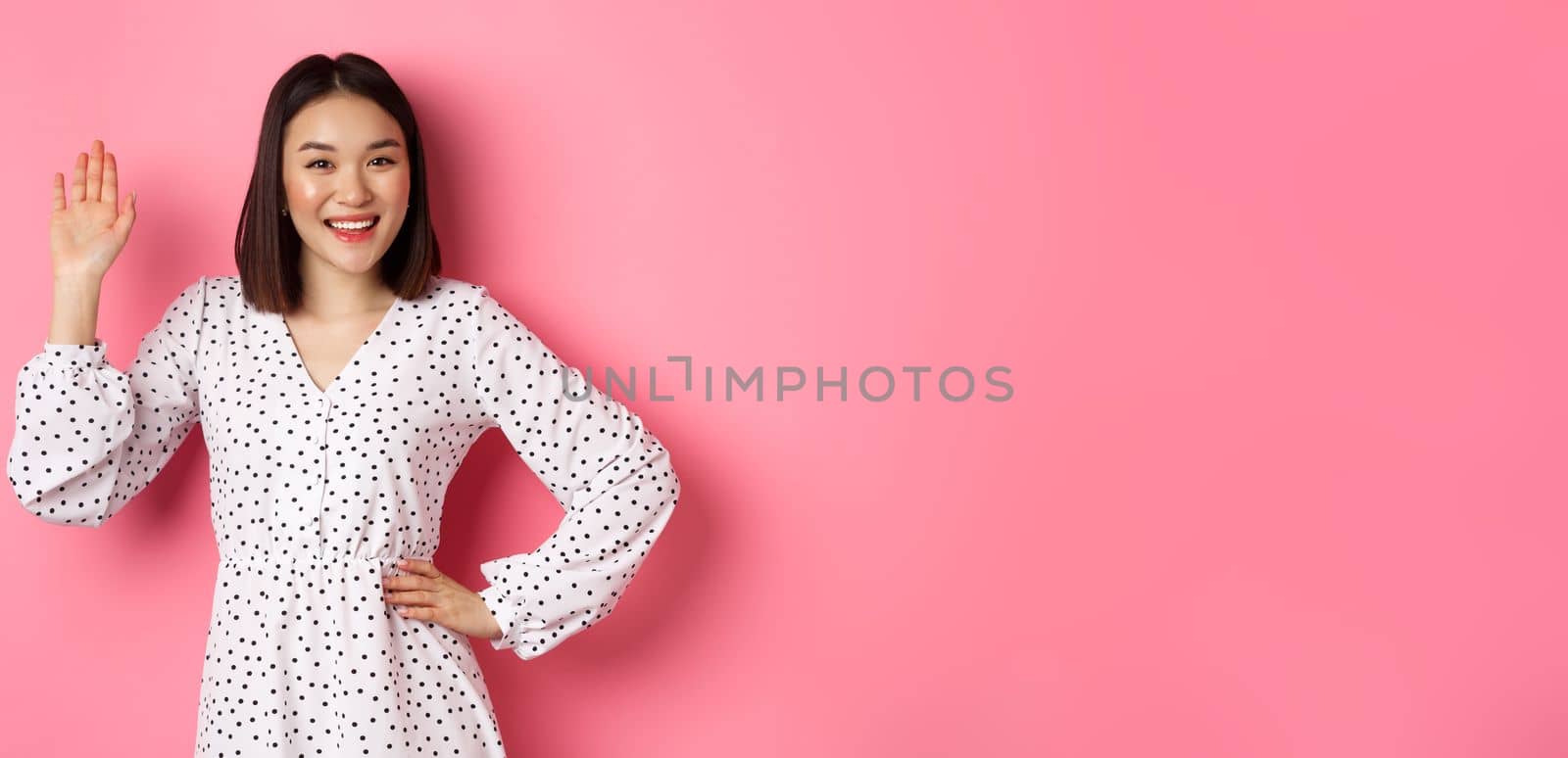 Beautiful asian woman in dress saying hello, waving hand to greet and say hi, smiling friendly at camera, standing over pink background.