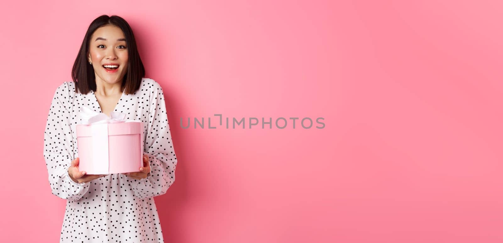 Surprised and happy asian woman receiving cute box with gift, standing amazed over pink background by Benzoix
