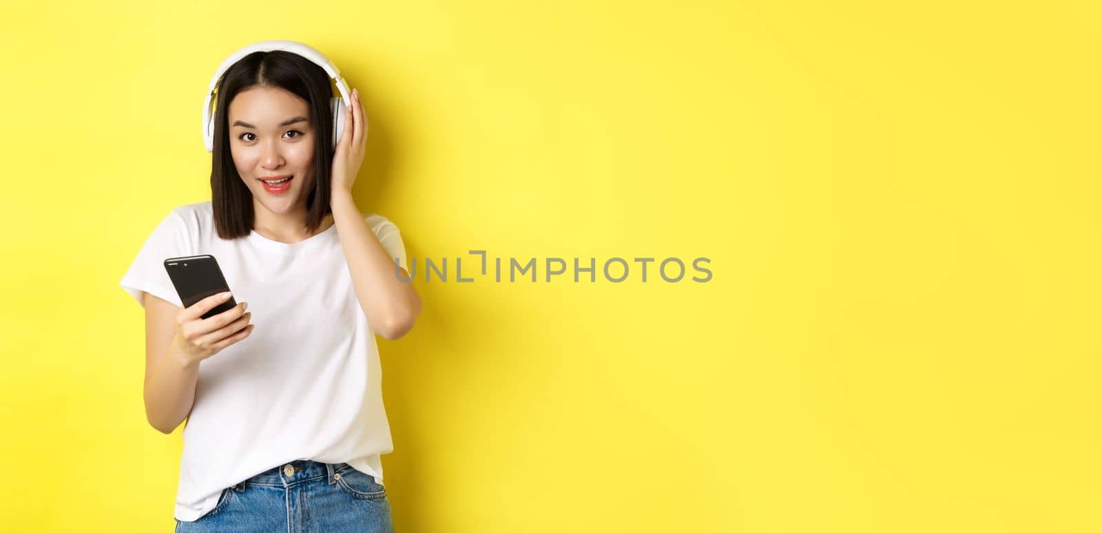 Modern asian woman listening music in wireless headphones, reading smartphone screen and smiling, standing in white t-shirt over yellow background by Benzoix