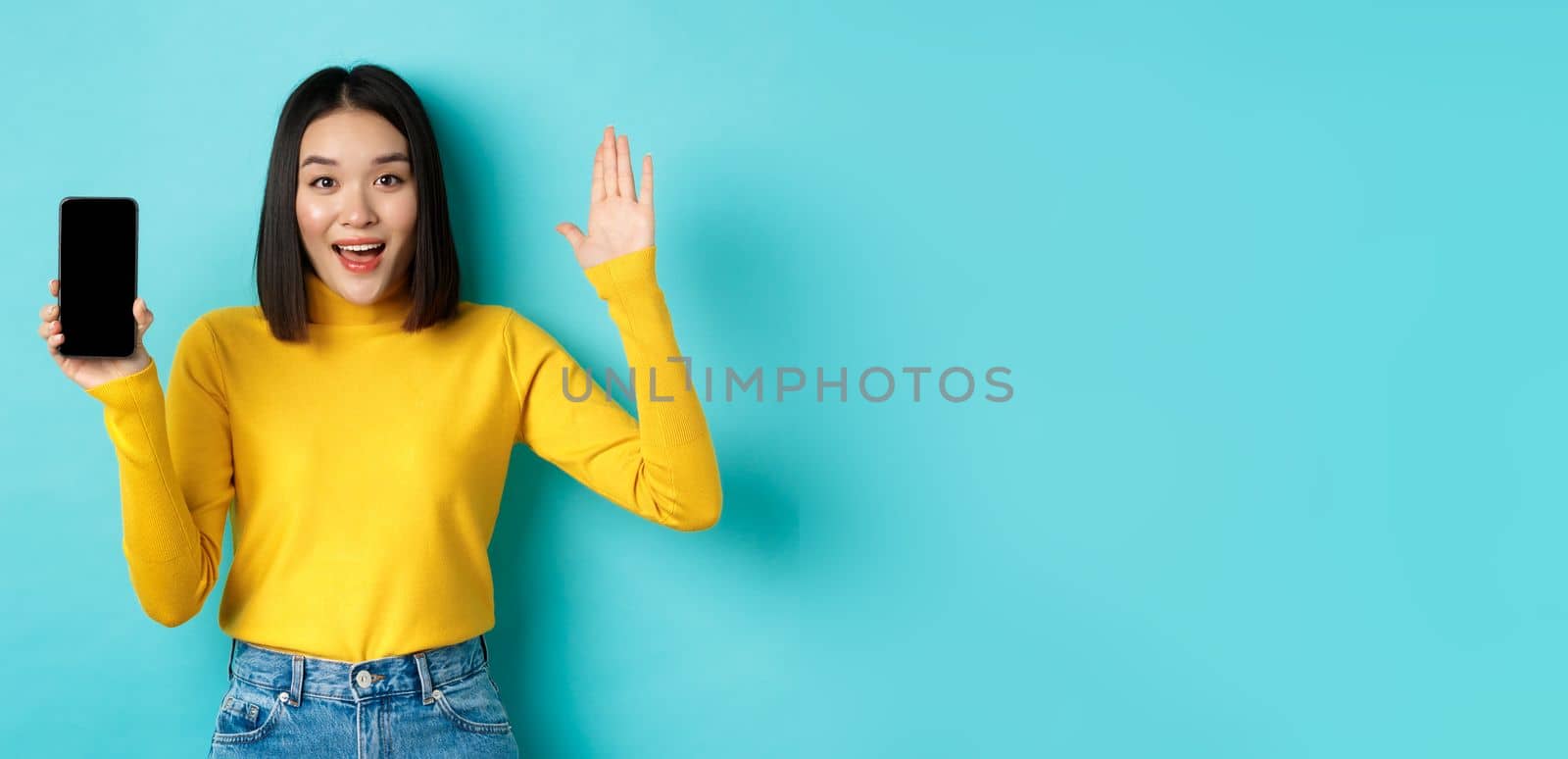 Cheerful asian woman showing empty smartphone screen and raising hand to say hello, greeting you, standing against blue background by Benzoix