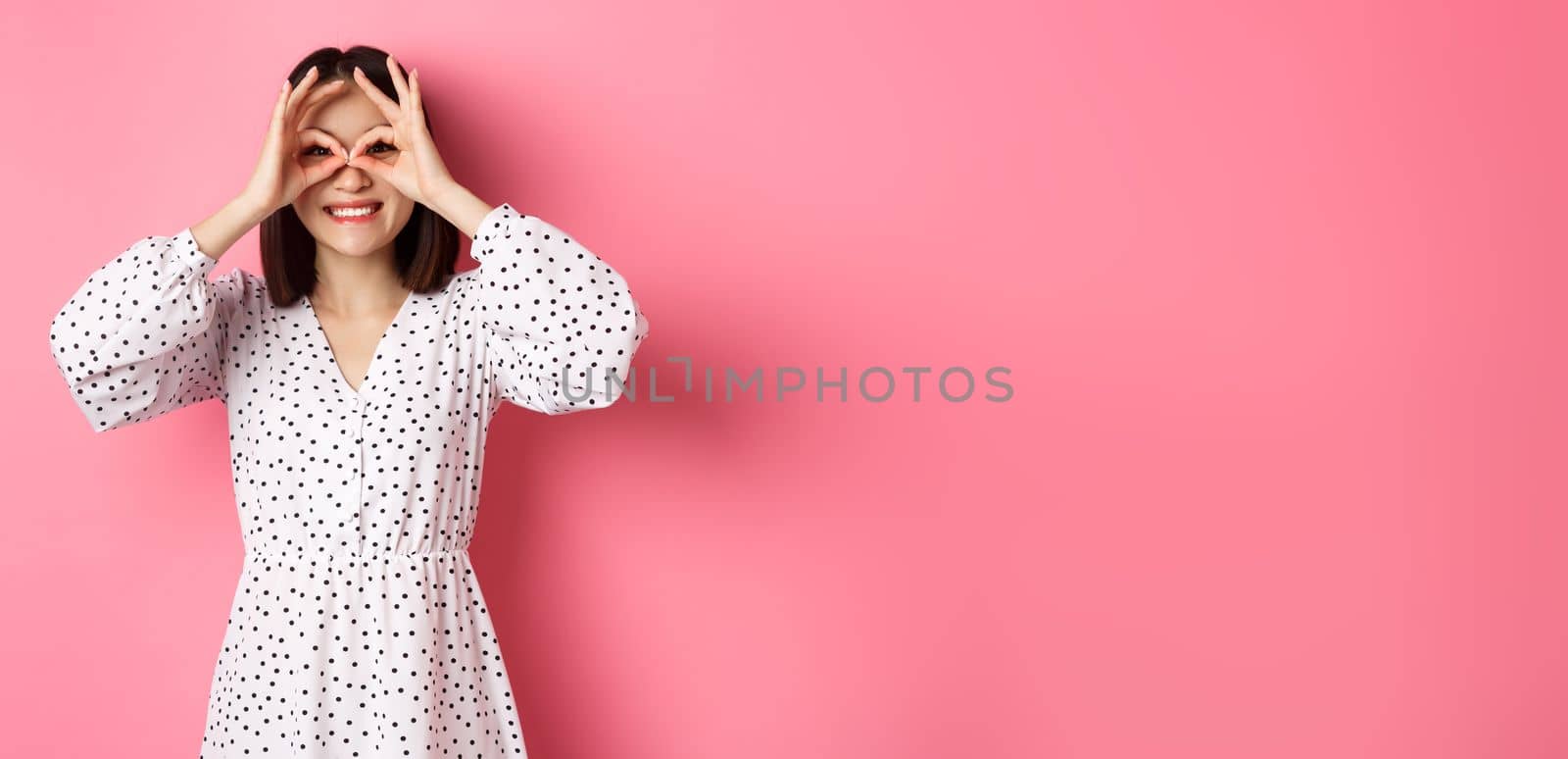 Carefree young asian woman looking through hand binoculars at camera, staring at discounts, standing over pink background by Benzoix