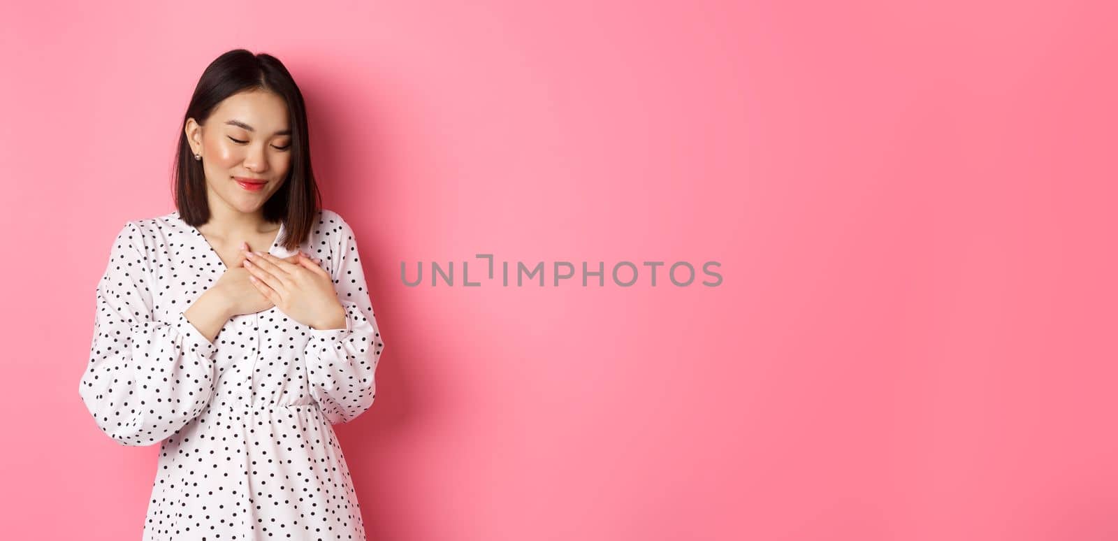 Beautiful korean girl in trendy spring dress dreaming, holding hands on heart, smiling with closed eyes, imaging something or having heartwarming memory, pink background.