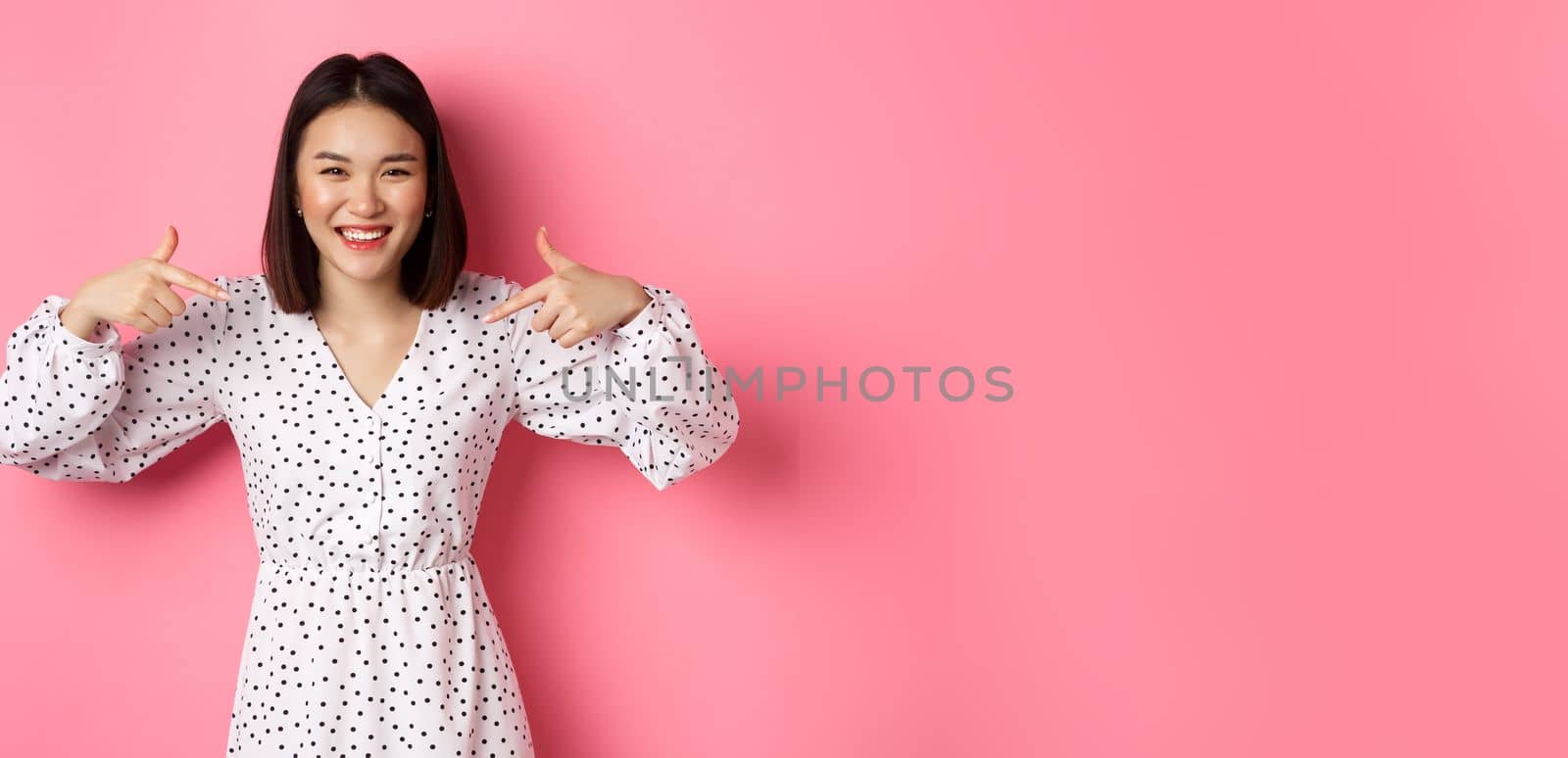 Beautiful korean woman pointing fingers at your logo and smiling, standing in dress over romantic pink background by Benzoix