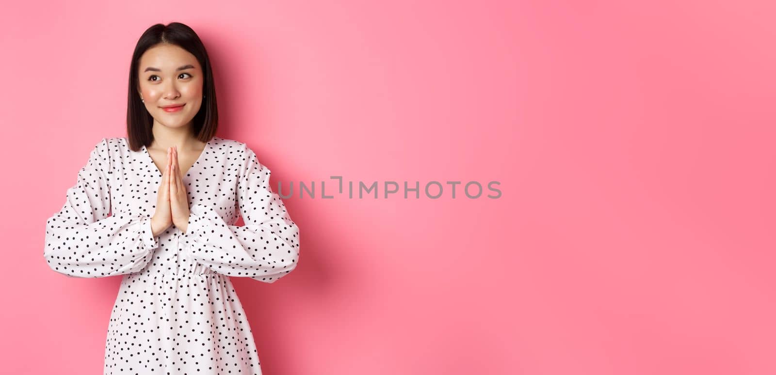 Beautiful angelic asian woman smiling, holding hands in pray and looking left at copy space with innocent cute gaze, standing over pink background.