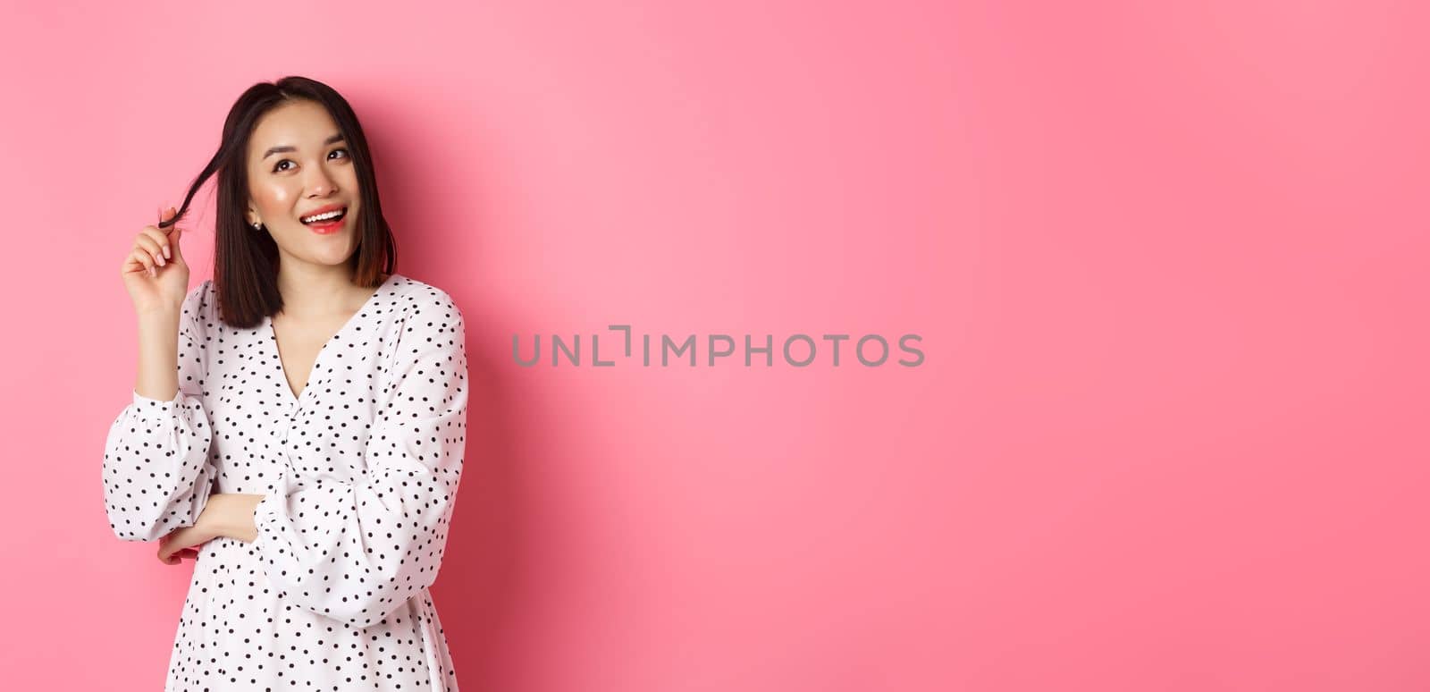 Dreamy cute asian girl thinking, imaging shopping and smiling, looking upper left corner with thoughtful smile, standing in dress over pink background by Benzoix