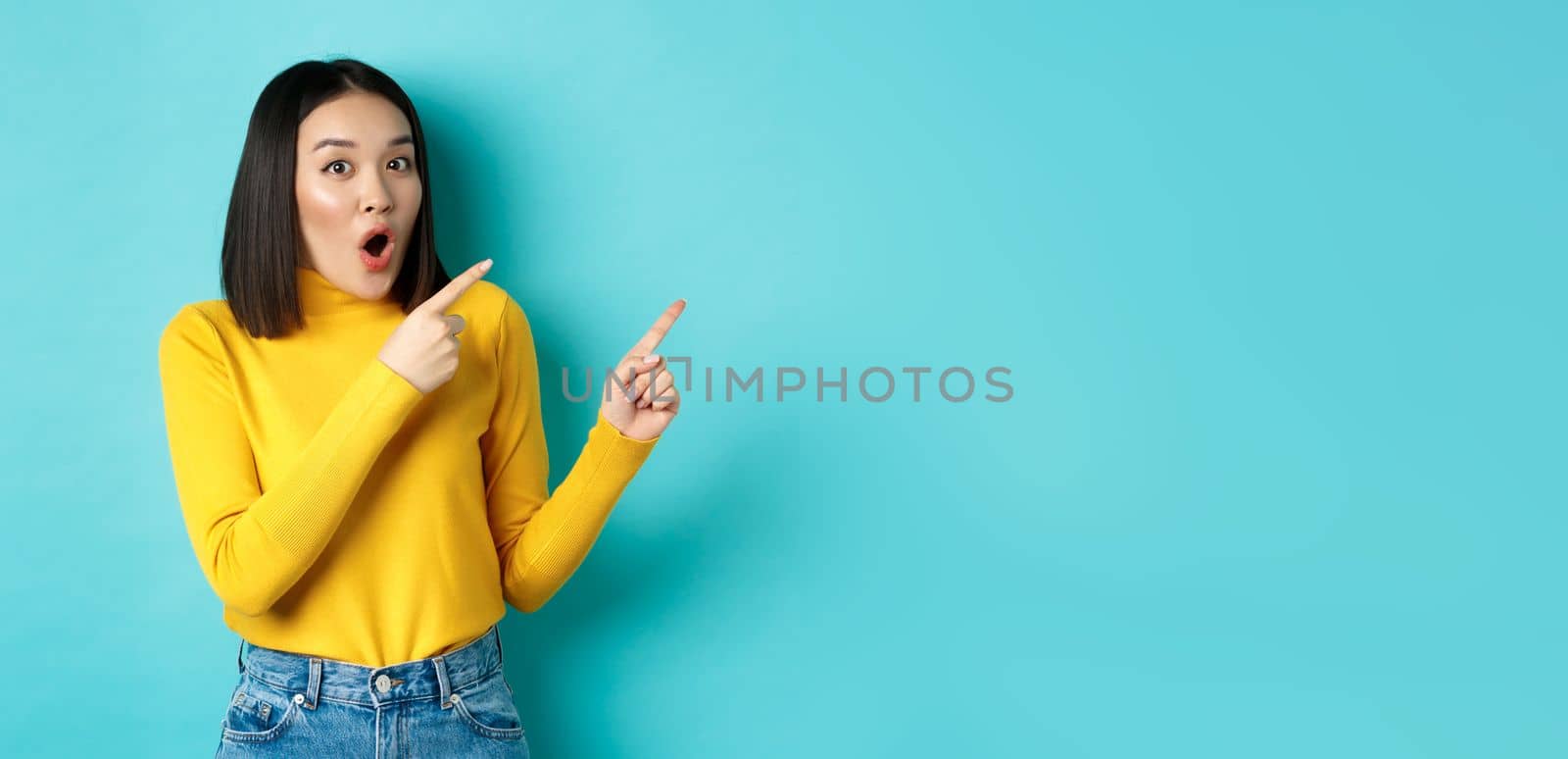 Shopping concept. Impressed korean girl say wow and pointing fingers at upper right corner, checking out special deal, standing over blue background by Benzoix
