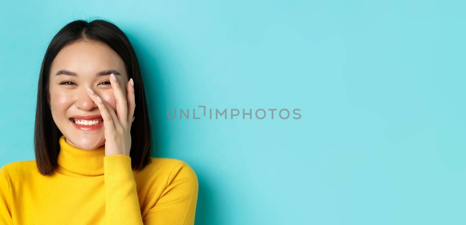 Beauty and makeup concept. Close up of beautiful asian woman blushing and laughing, touching glowing healthy skin, smiling happy, standing over blue background.