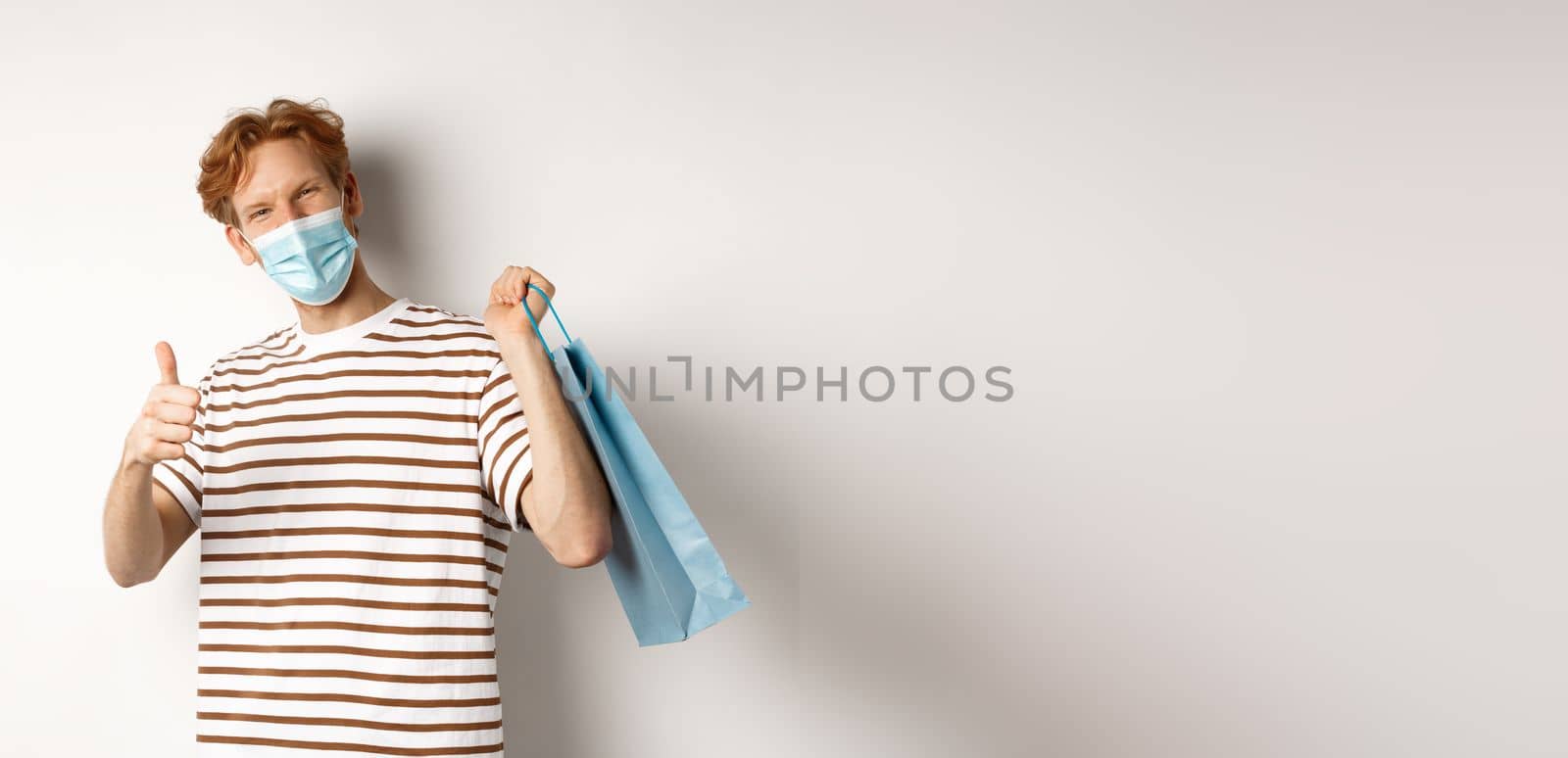Concept of covid-19 and shopping. Satisfied young man looking pleased after shopping, wearing face mask, showing thumbs-up, recommend store, white background.