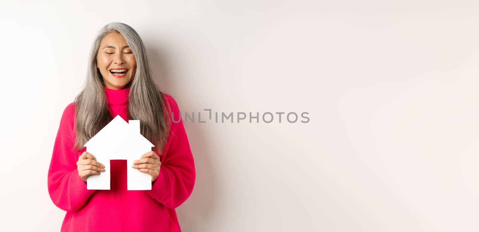 Real estate. Happy asian grandmother laughing with eyes closed, holding paper house model, dreaming about paper house model, standing over white background. Copy space