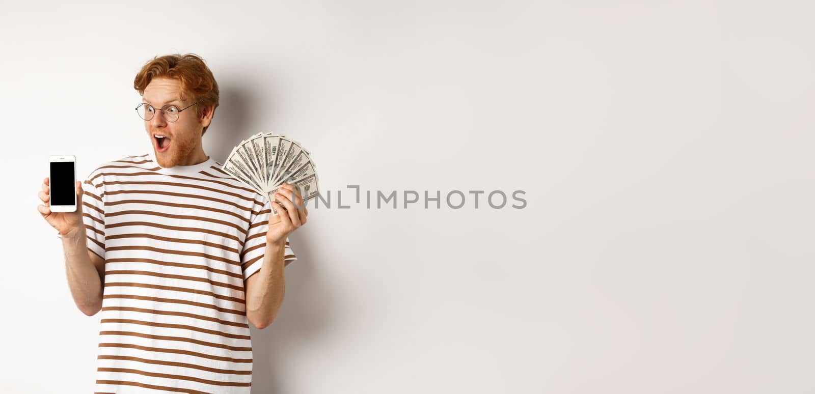 Amazed redhead man showing smartphone app on blank screen and money, winning prize cash online, standing over white background by Benzoix