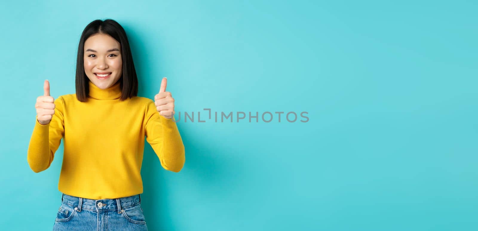 Cheerful asian female model showing thumbs up gesture, smiling and looking impressed, praise good product, standing over blue background by Benzoix