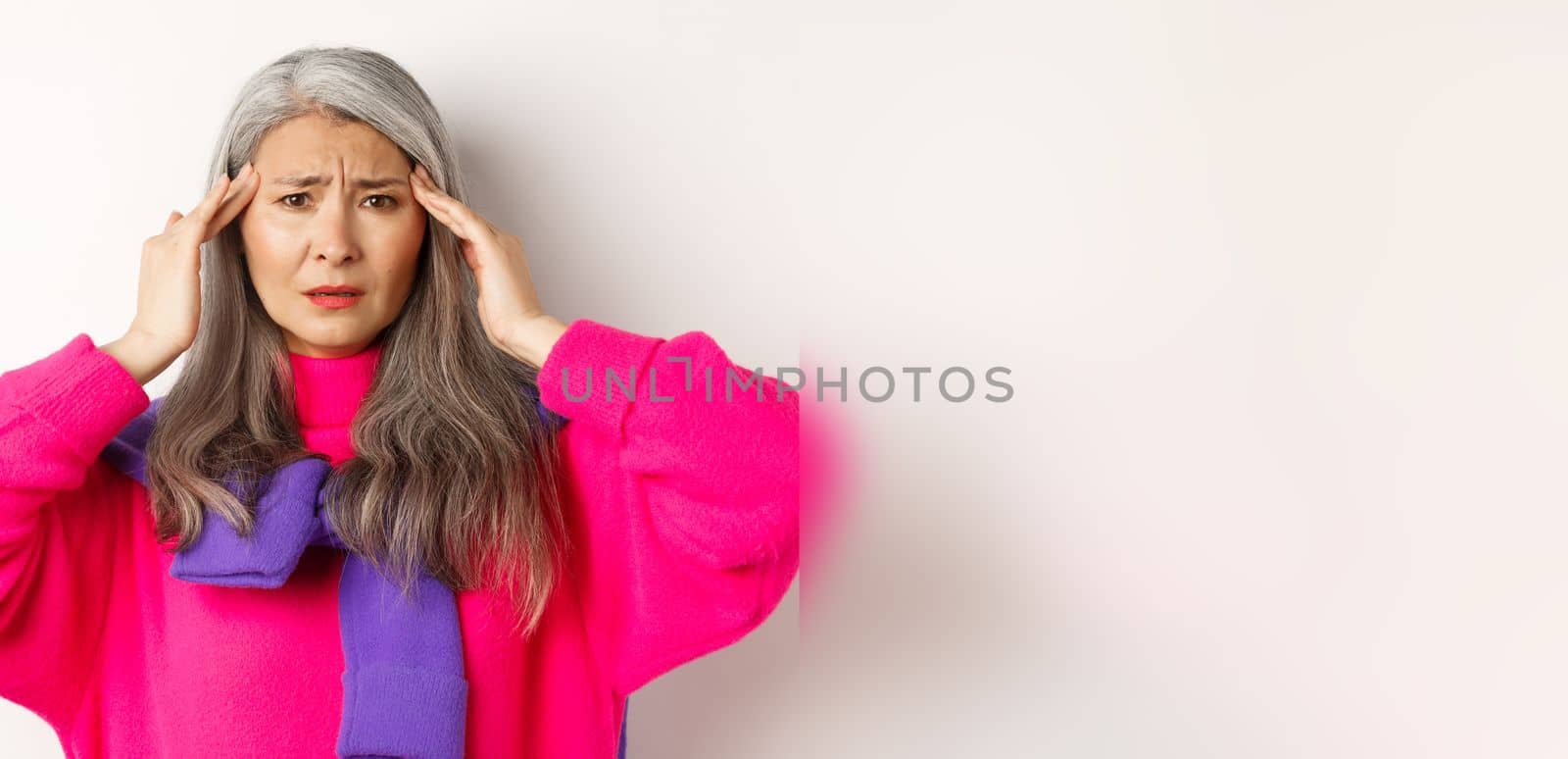 Close-up of fashionable asian senior woman feeling unwell, touching head and grimacing from headache, complain on migraine, white background.
