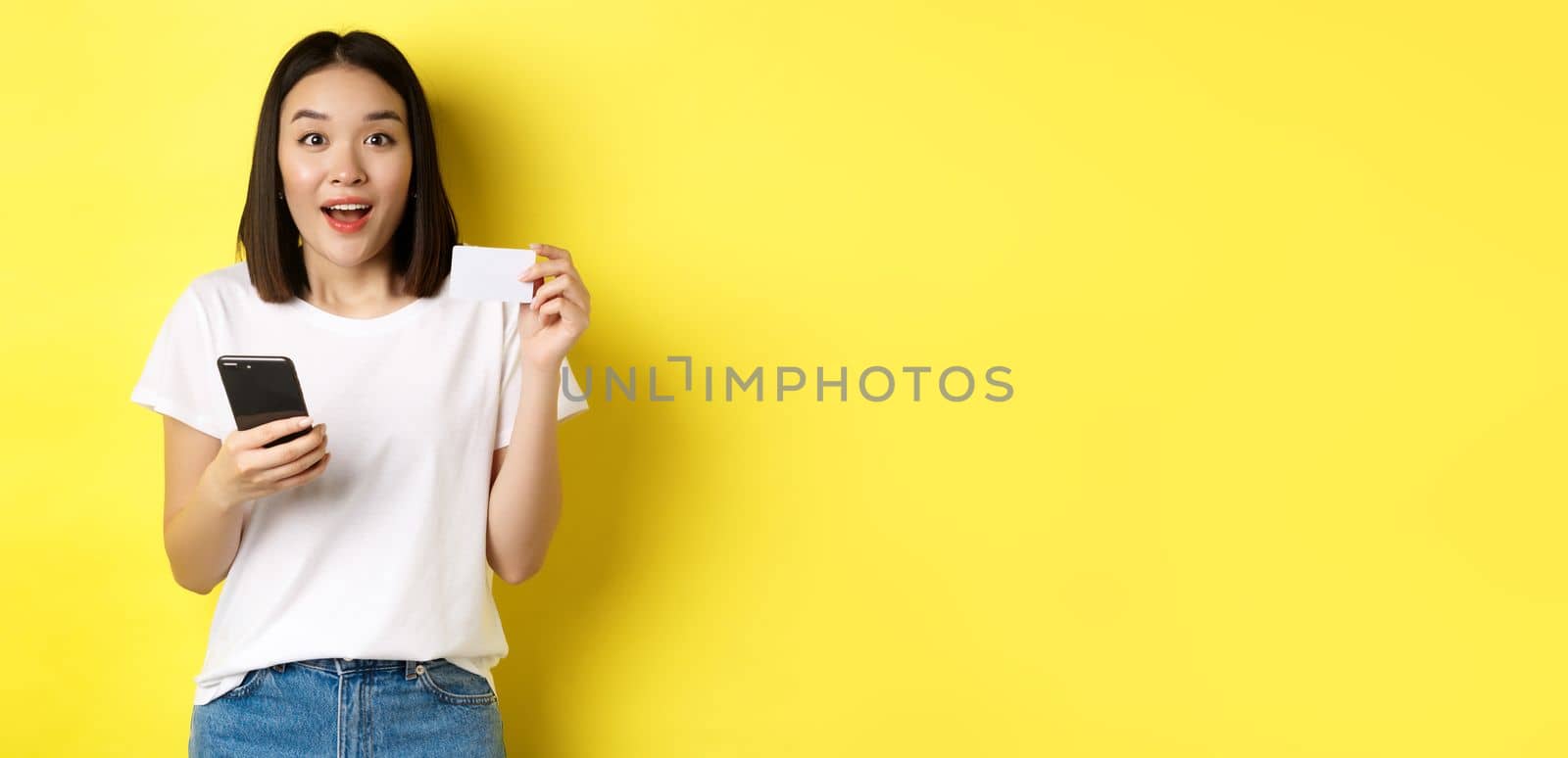E-commerce and online shopping concept. Excited asian woman order in internet, holding smartphone and plastic credit card, standing over yellow background.