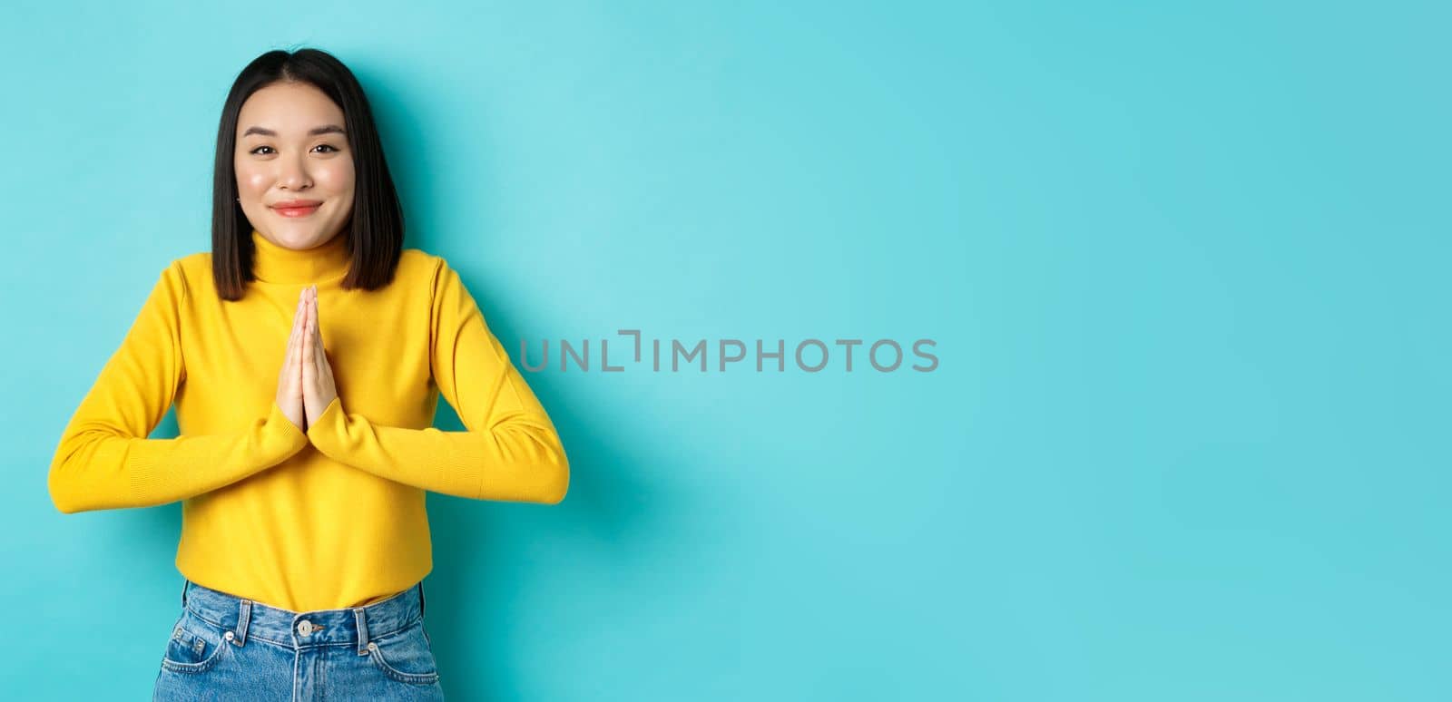 Cute asian woman in trendy outfit saying thank you, holding hands in namaste, begging gesture, smiling grateful at camera, standing over blue background.