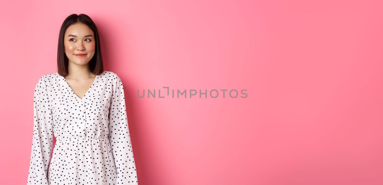 Cute asian woman smiling, looking left at copy space, standing on romantic pink background by Benzoix