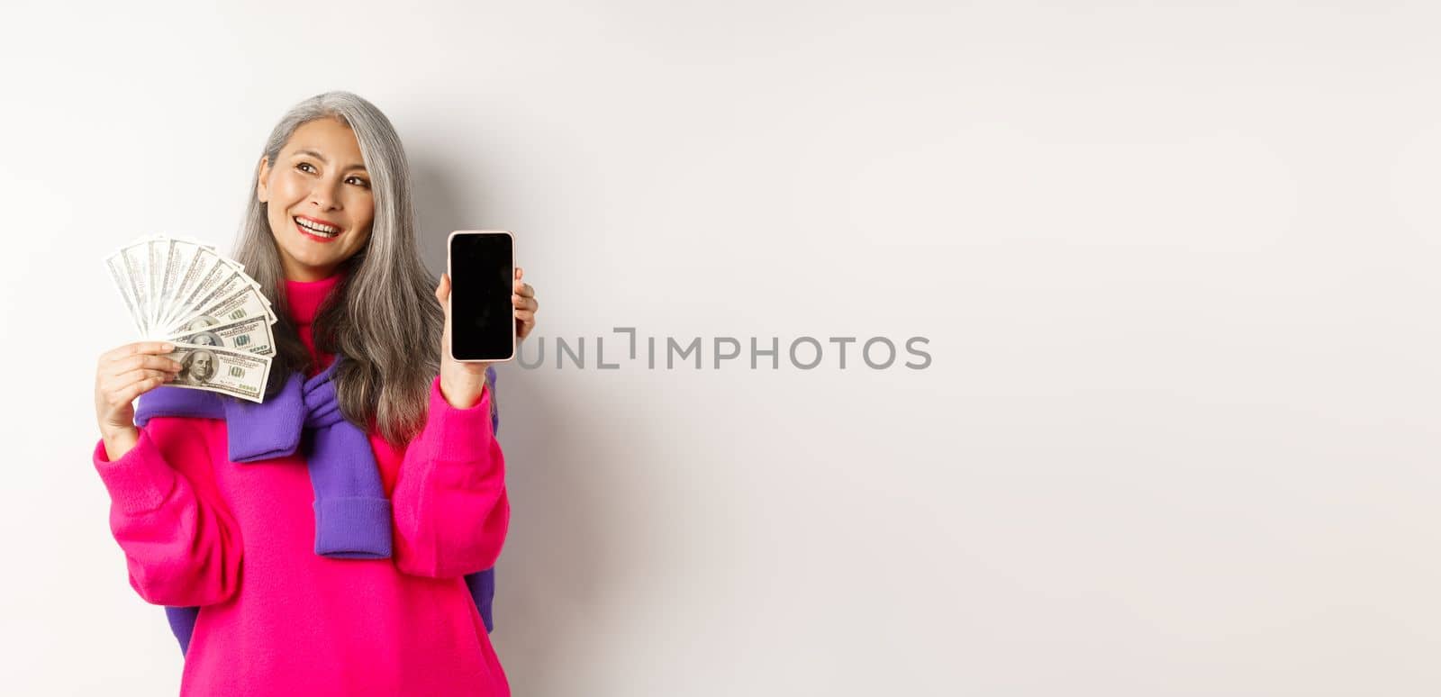 Fashionable asian senior woman showing money dollars and blank smartphone screen, demonstrate online shop, standing over white background by Benzoix