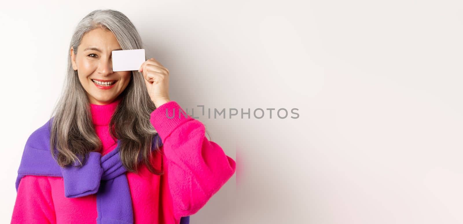 Shopping concept. Stylish asian senior woman smiling and showing plastic credit card, paying contactless, standing over white background by Benzoix