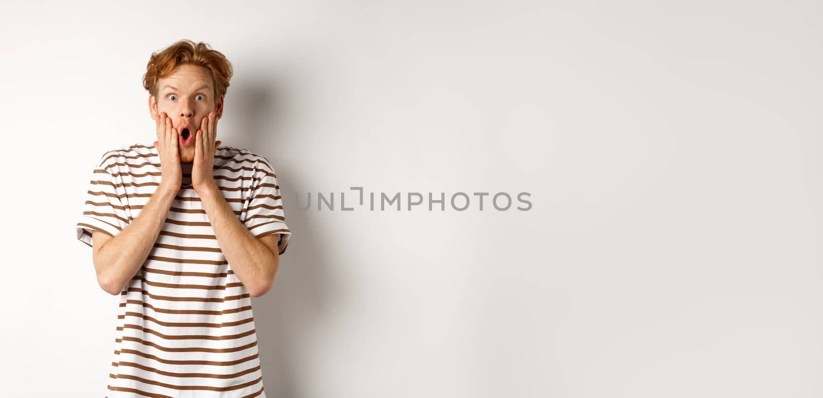 Impressed young man with ginger hair, gasping and staring shocked at camera, express complete disbelief and amazement, white background.