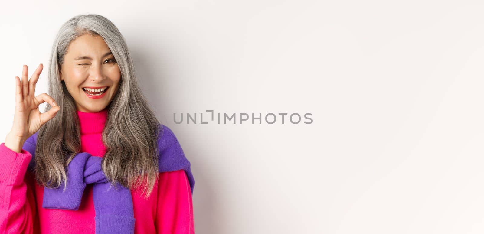 Close-up of cheerful asian grandmother in fashionable pink sweater, winking and showing okay sign, recommending promotion, standing over white background by Benzoix