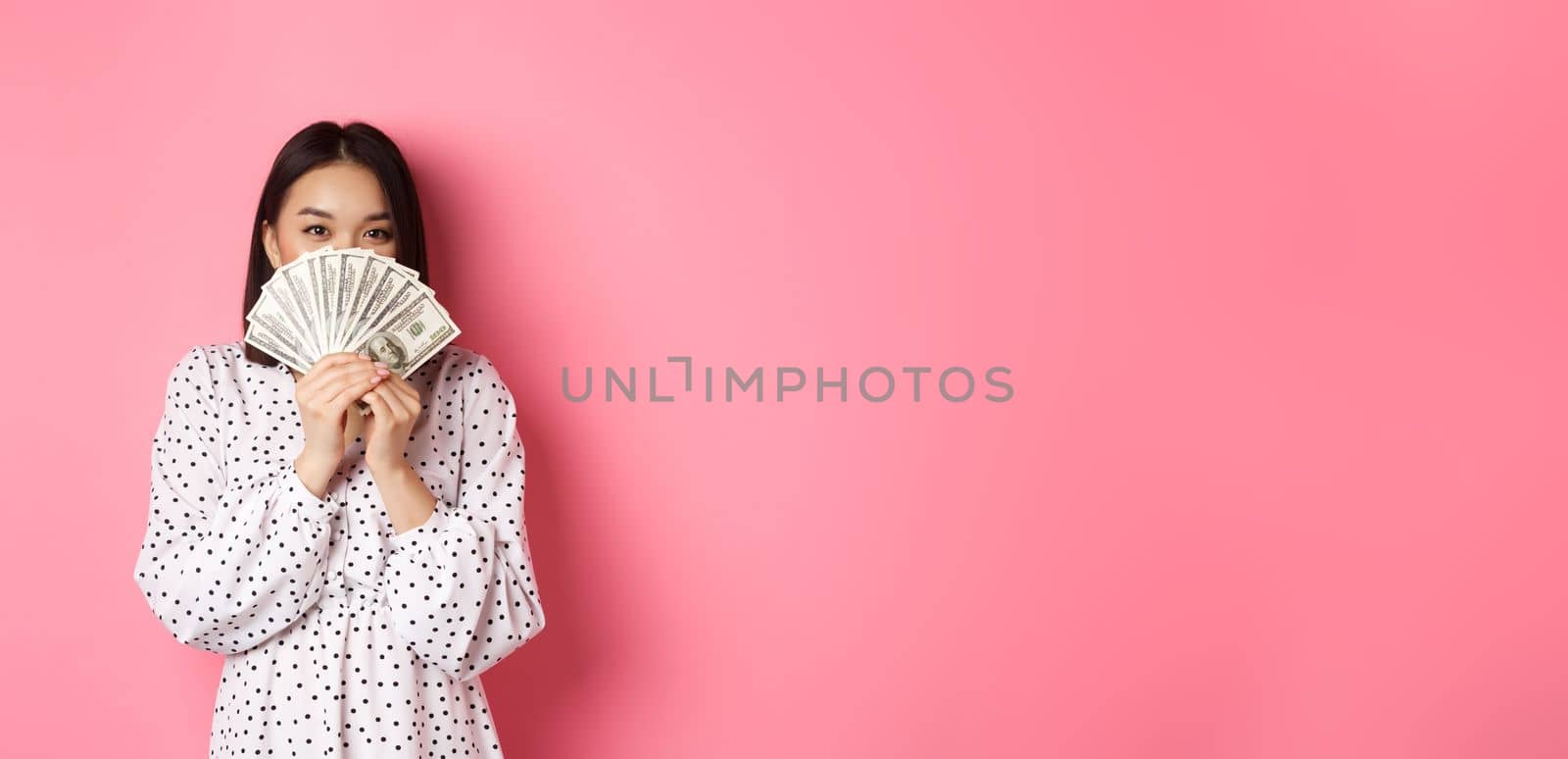 Shopping concept. Cute asian woman hiding face behind money dollars, peeking at camera, standing over pink background.