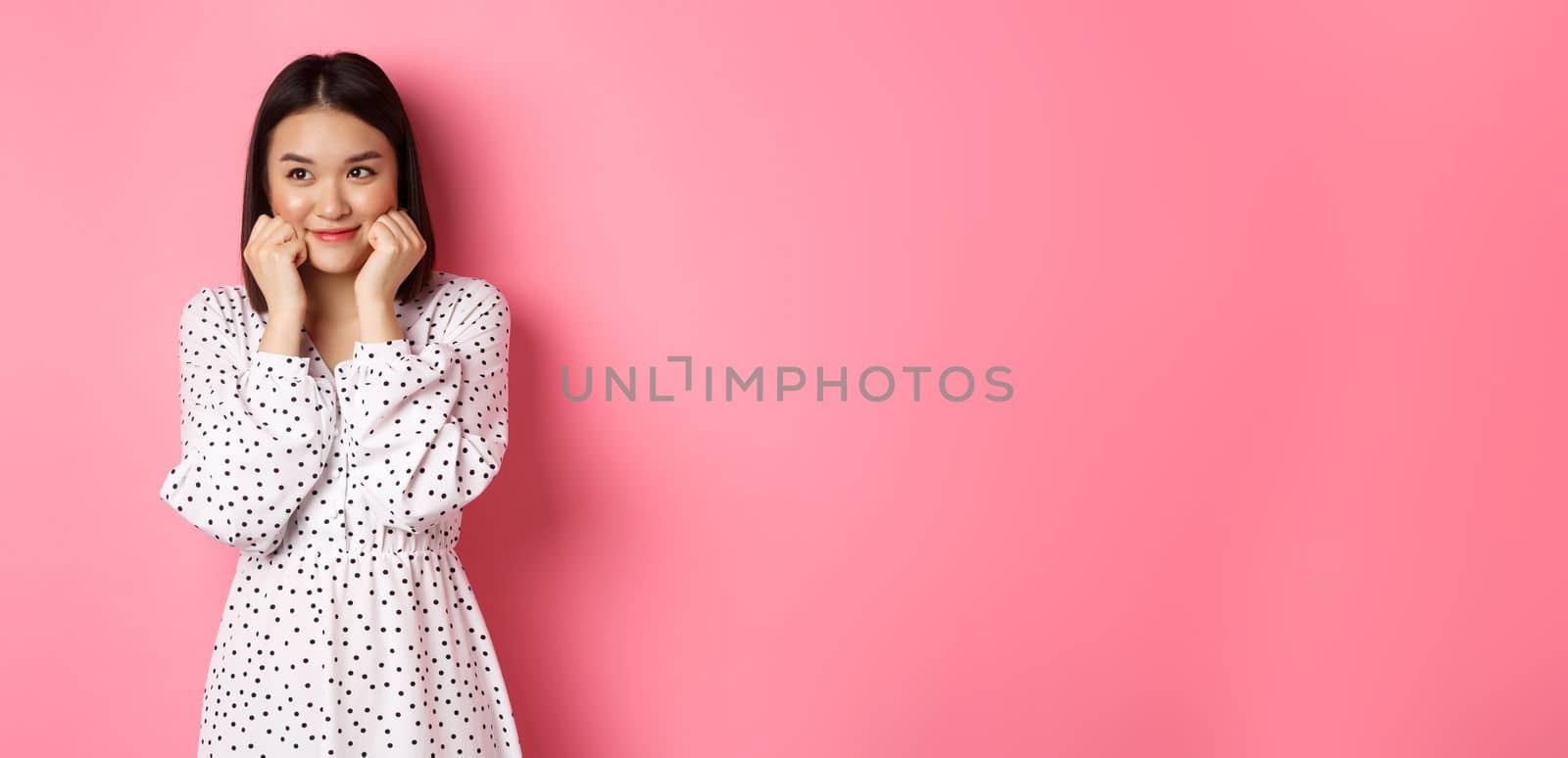 Cute and shy asian girl blushing, touching cheeks and looking left at copy space silly, standing against pink background.