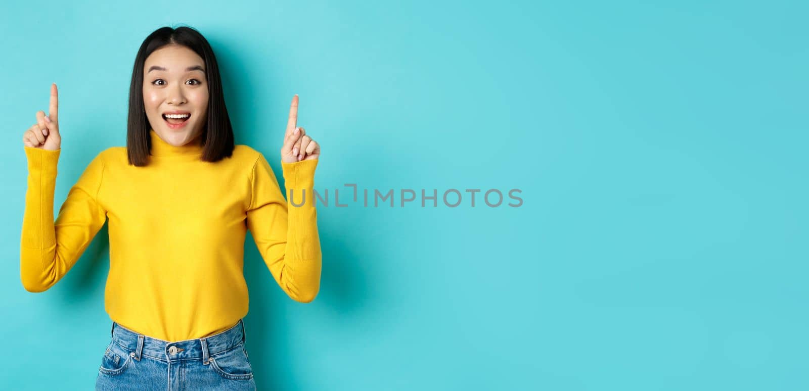 Shopping concept. Impressed young korean woman showing advertisement on top, pointing fingers up and gasping amazed, saying wow, standing over blue background.