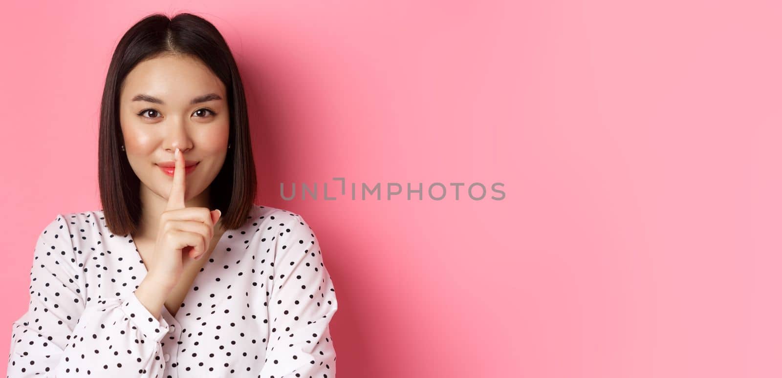 Close-up of mysterious asian woman hiding a secret, hushing and telling to keep quiet, standing over pink background by Benzoix