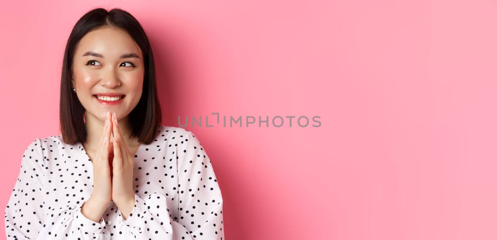 Beauty and lifestyle concept. Close-up of happy and satisfied asian woman clap hands, expect something good, smiling and looking aside, standing over pink background.