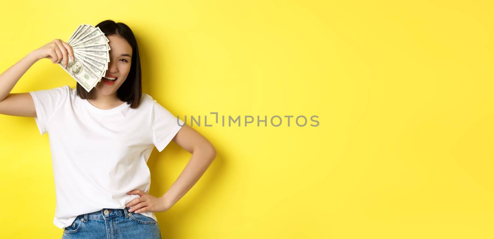 Cute asian woman hiding face behind money, peeking at camera satisfied, earn cash, standing over yellow background by Benzoix