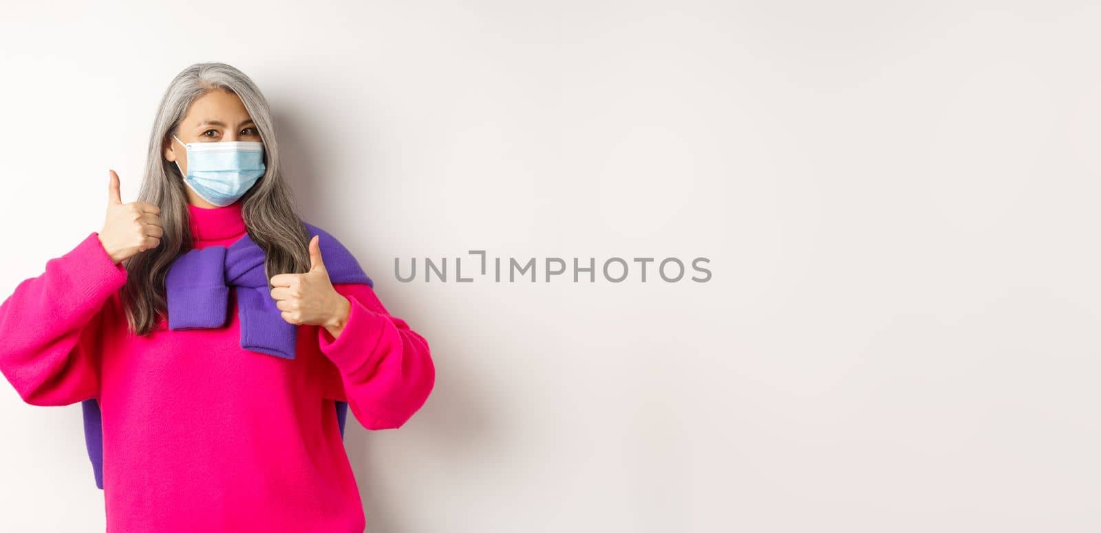 Covid, pandemic and social distancing concept. Cheerful and stylish asian senior woman wearing medical mask and showing thumbs-up, standing over white background.