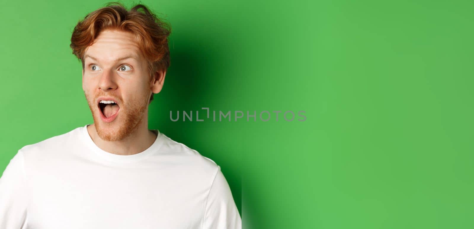 Close up of surprised and impressed redhead man checking out promotion offer, looking left with dropped jaw, standing over green background.