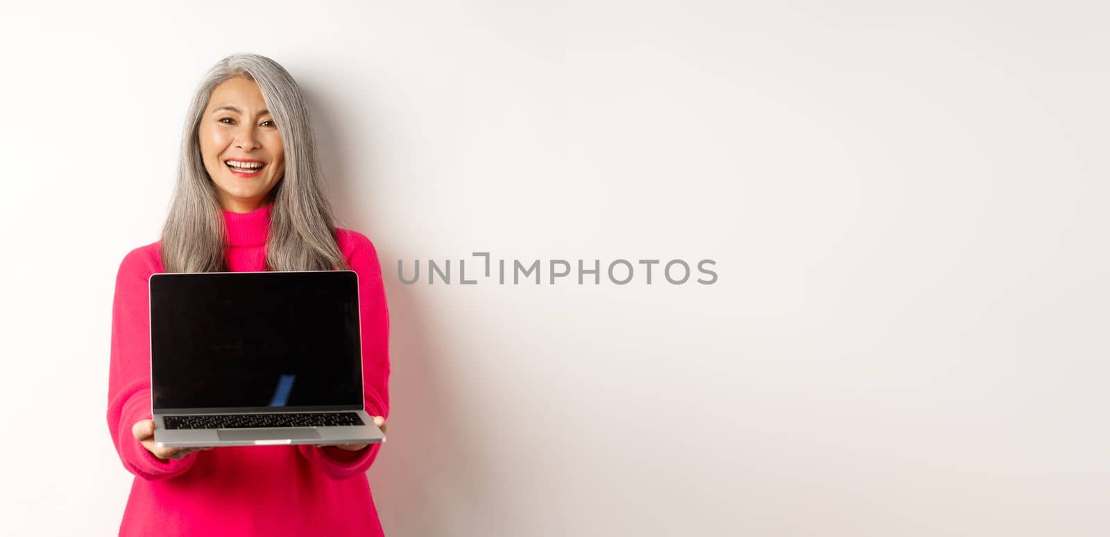 E-commerce concept. Beautiful asian senior businesswoman showing blank laptop screen and smiling, standing in pink sweater over white background.