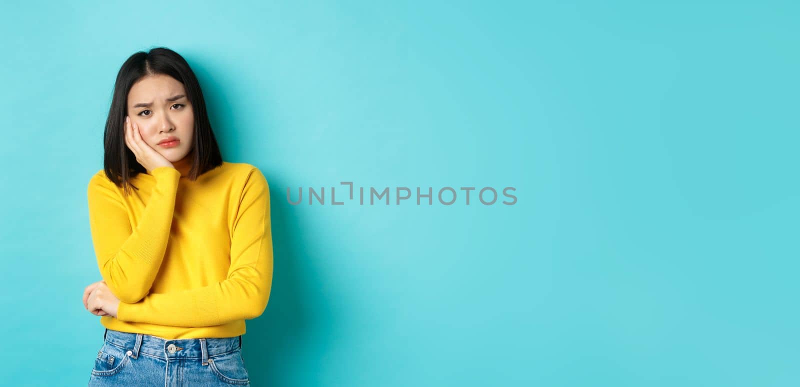 Sad and bored asian girl looking reluctant and unamused at camera, leaning face on hand, standing over blue background.