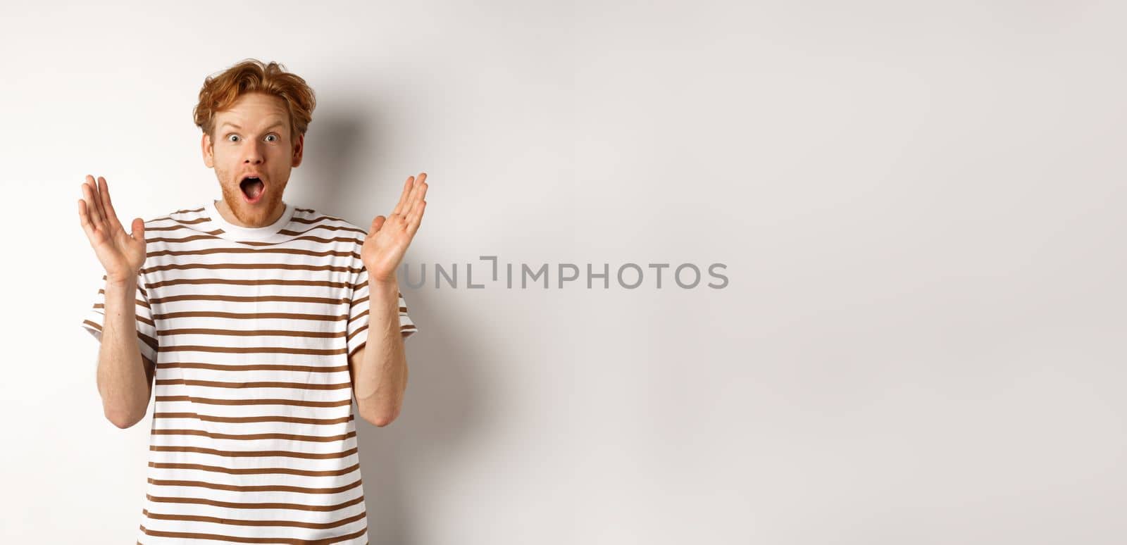 Surprised redhead man scream of amazement, staring with disbelief and excitement, hear awesome news, standing over white background.