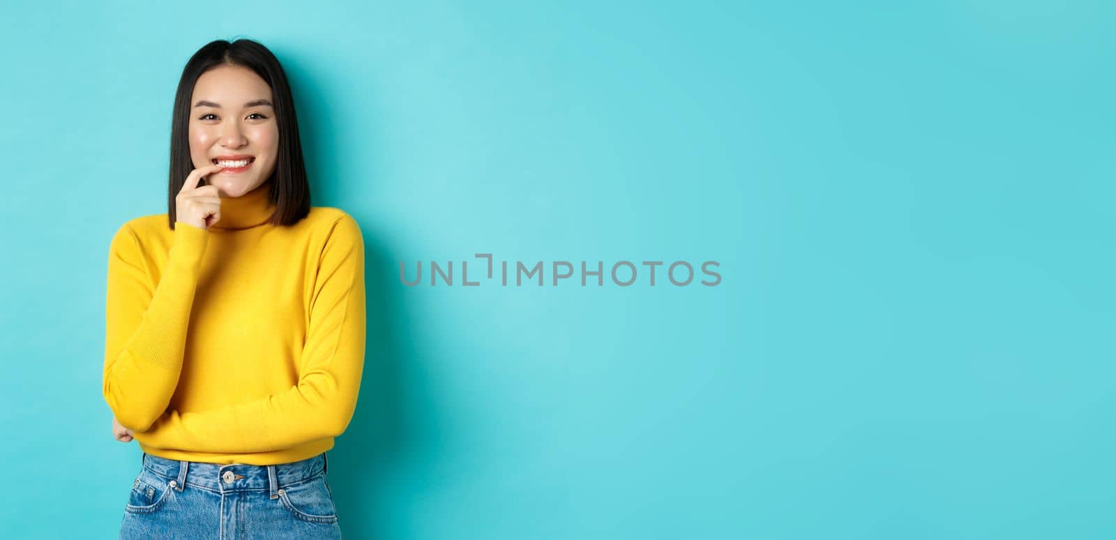 Image of good-looking asian woman in stylish outfit, touching lip and smiling at camera with happy face, standing over blue background by Benzoix
