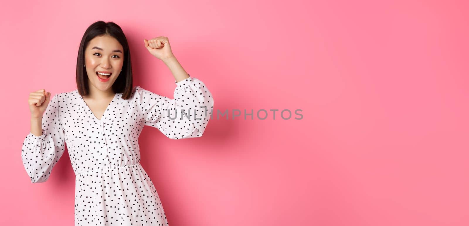 Beautiful korean woman dancing and having fun, smiling happy at camera, posing against pink background.