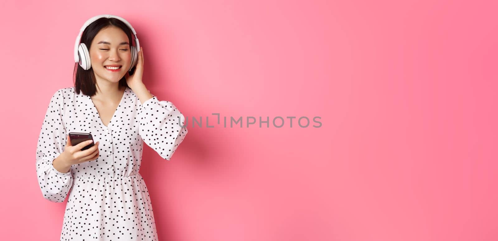 Romantic asian girl listening music in headphones, smiling with closed eyes, holding mobile phone, standing over pink background by Benzoix