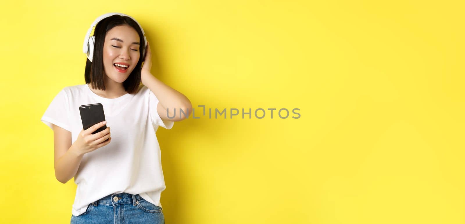 Cool asian girl singing along and listening music in wireless headphones, holding smartphone in hand, standing over yellow background by Benzoix