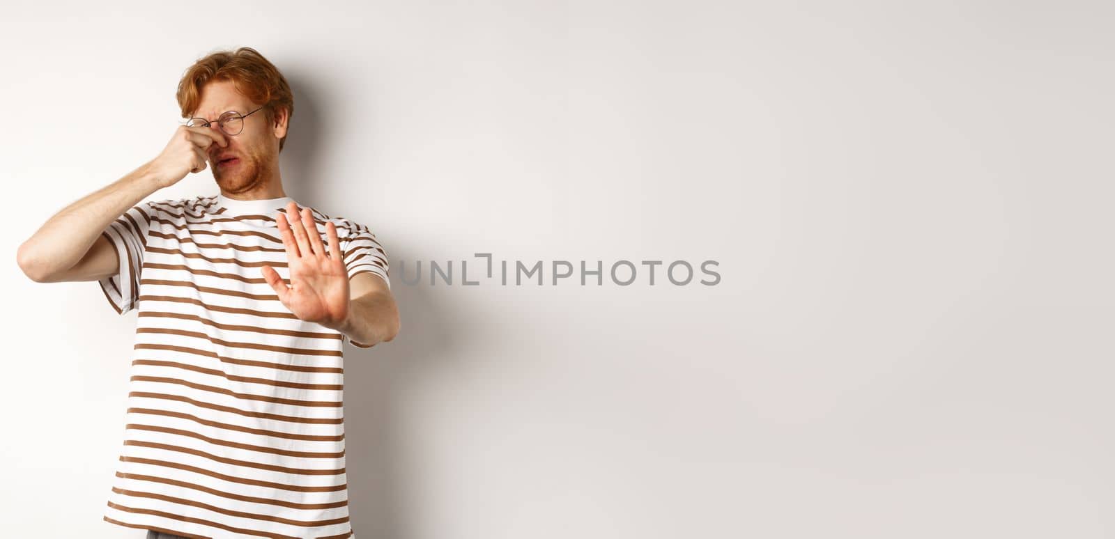 Young man with red hair staring at camera and shut nose from bad smell, looking disgusted and refusing, white background.