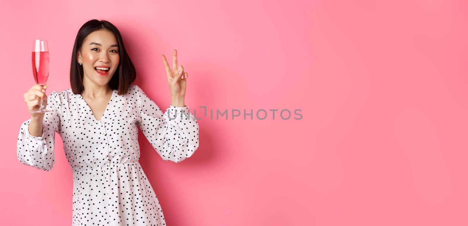 Cute asian female model drinking champagne, celebrating on party and showing peace sign, smiling happy at camera, standing over pink background by Benzoix