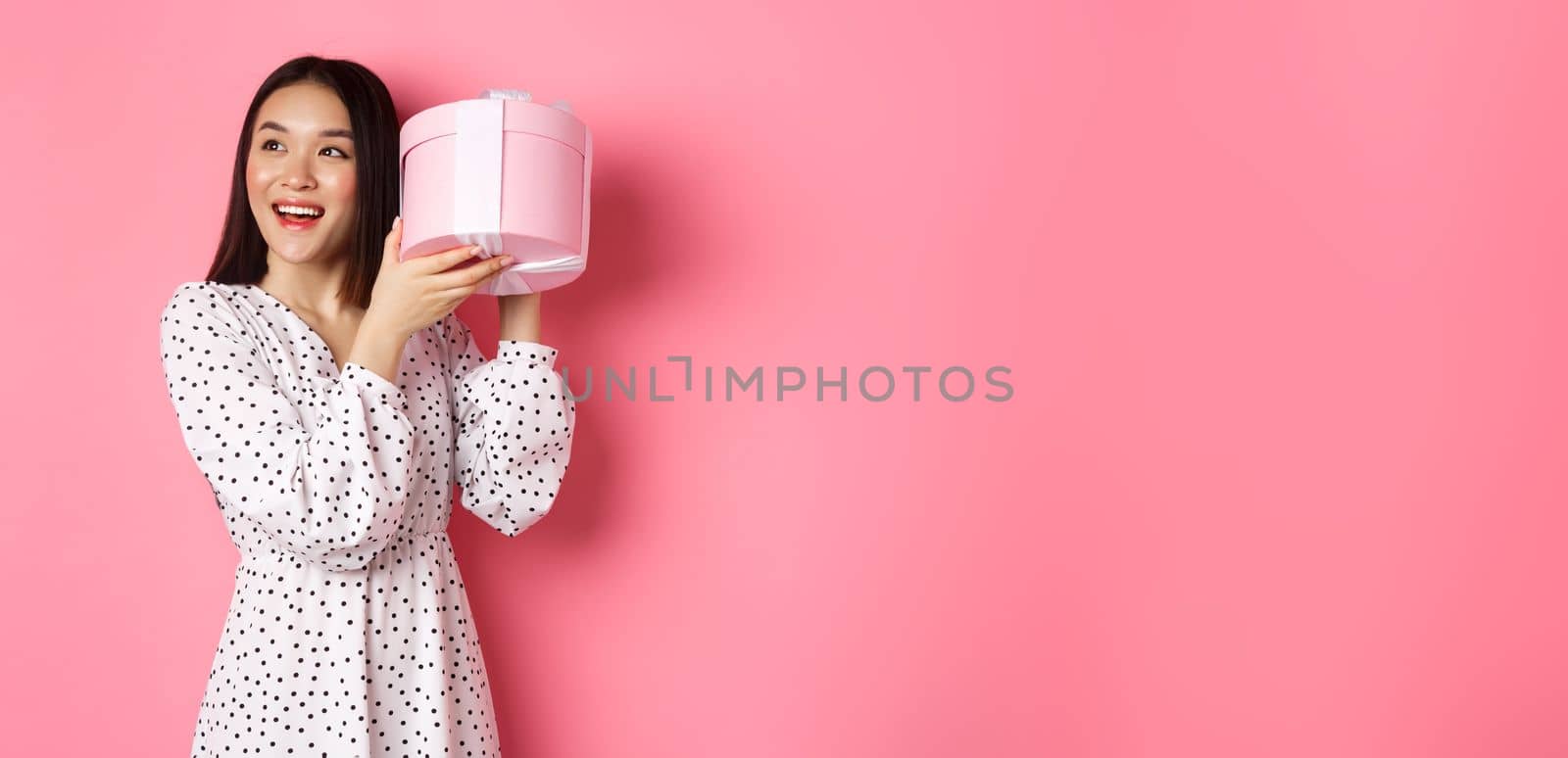 Cute asian woman shaking box with gift, smiling and looking intrigued, guess what inside present, standing over pink background by Benzoix